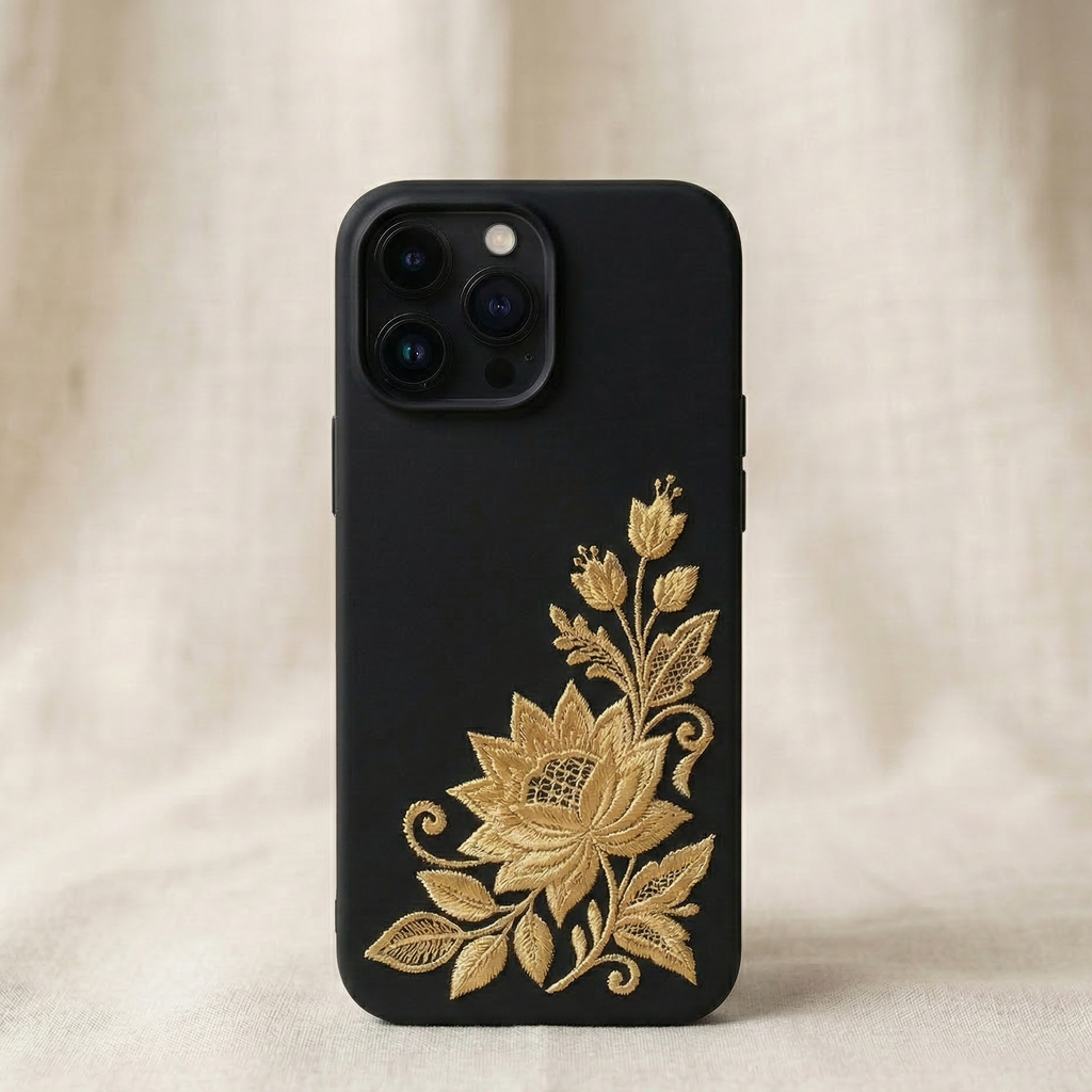 Minimalist luxury iPhone silicone phone case with black matte background featuring a small delicate gold embroidered jasmine flower in the bottom right corner, inspired by traditional South Asian embroidery, fine white thread stitching with realistic raised embroidered texture, elegant minimal composition with lots of negative space