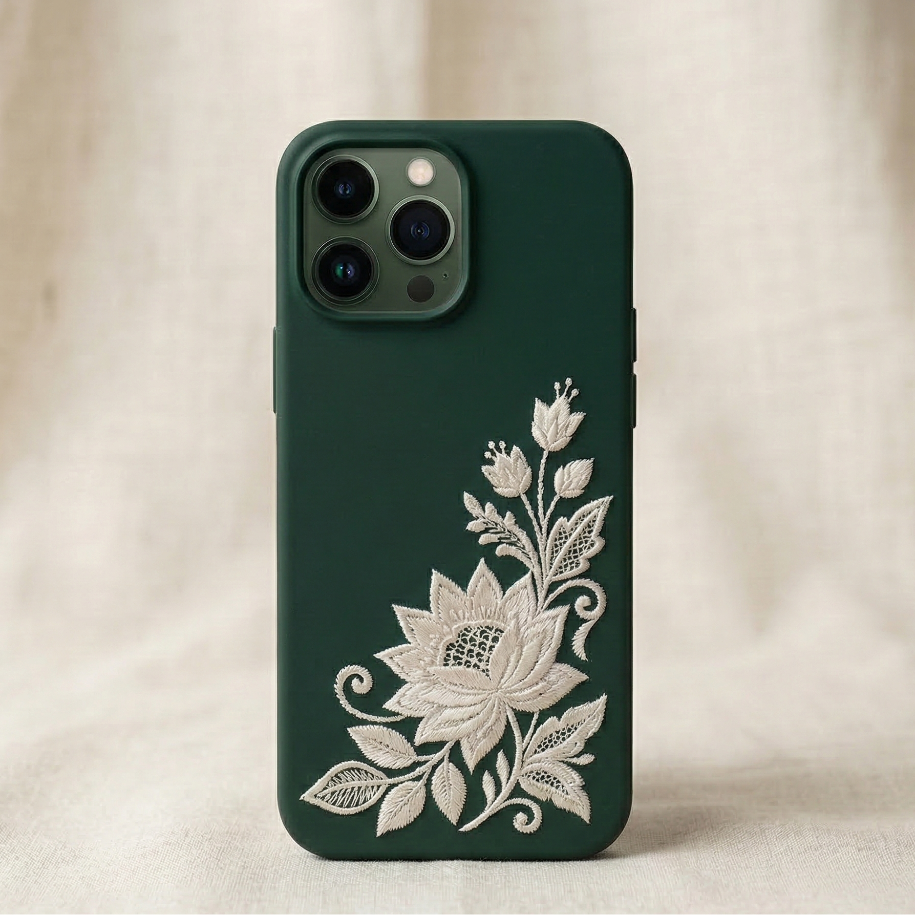 Minimalist luxury iPhone silicone phone case with deep emerald green matte background featuring a small delicate embroidered jasmine flower in the bottom corner, inspired by traditional South Asian embroidery, fine white thread stitching with realistic raised embroidered texture, elegant minimal composition with lots of negative space, refined modern South Asian luxury aesthetic