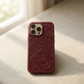 Luxury maroon embossed iPhone case featuring intricate South Asian floral and paisley patterns inspired by traditional Pakistani naqashi and textile motifs, deep maroon leather texture with detailed raised mandala carving, photographed standing upright on a soft neutral fabric surface with warm natural sunlight coming from a window.